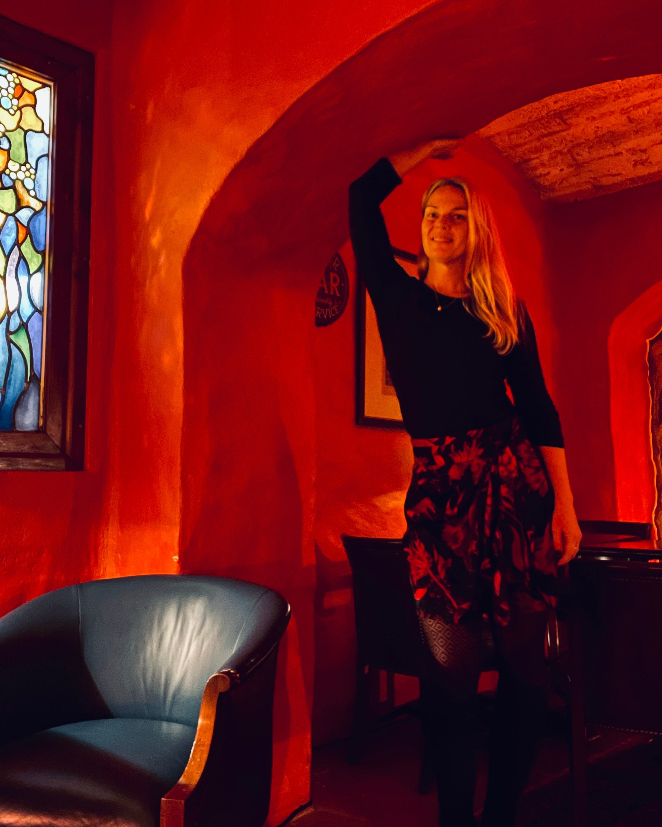 Woman in a black top and floral skirt standing in a warmly lit room with a stained glass window.